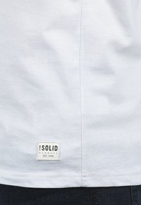 Hvid bomulds t-shirt med et rektangulært mærke med sort tekst, placeret nederst. Søm detaljer synlige langs siden.