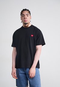 Zwart katoenen T-shirt met een klein rood hartlogo op de linkerkant van de borst. Gepaard met blauwe denim shorts, die een ontspannen pasvorm tonen.