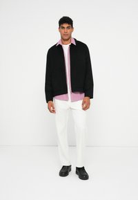 Mann iført svart jakke over rosa skjorte, hvite bukser og svarte sko, står mot en enkel hvit bakgrunn.