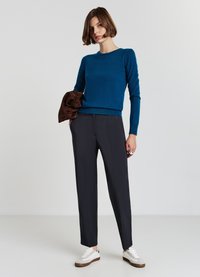 Blauer Strickpullover mit langen Ärmeln, kombiniert mit grauen maßgeschneiderten Hosen. Das Modell hält eine braune Jacke und trägt weiße Schuhe mit braunen Akzenten.