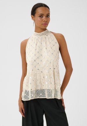 Femme portant un haut crème sans manches à col licou avec des sequins argentés et un pantalon noir, debout devant un fond clair uni.