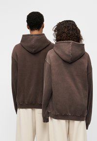 Καφέ φούτερ με κουκούλα σε χαλαρή εφαρμογή, με κορδόνι στη κουκούλα και ριμπ μανσέτες, συνδυασμένα με παντελόνια σε κρεμ χρώμα.