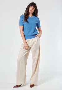 Sininen lyhythihainen neulepusero, jossa on teksturoitu timanttikuvio, yhdistettynä korkeavyötäröisiin vaaleanpunoisiin leveälahkeisiin housuihin ja tummanpunaisiin korkokenkiin.