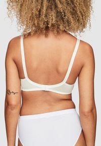 Vitt bh med justerbara band, slät yta och knäppning. Ett par vita trosor syns under; hudtonen är neutral.