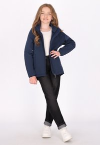 Mørkeblå softshell-jakke med høj krave, lynlås foran, sidelommer, båret over en hvid skjorte, parret med sorte bukser med opsmøg og hvide sneakers.