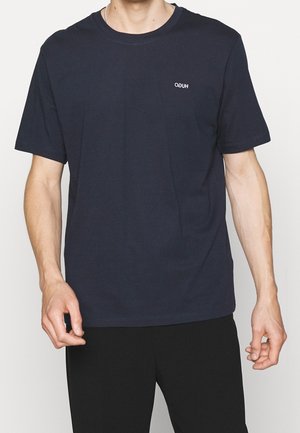 Mand iført en ensfarvet mørkeblå t-shirt med korte ærmer og et lille hvidt "HUGO"-logo på venstre bryst, kombineret med sorte bukser.