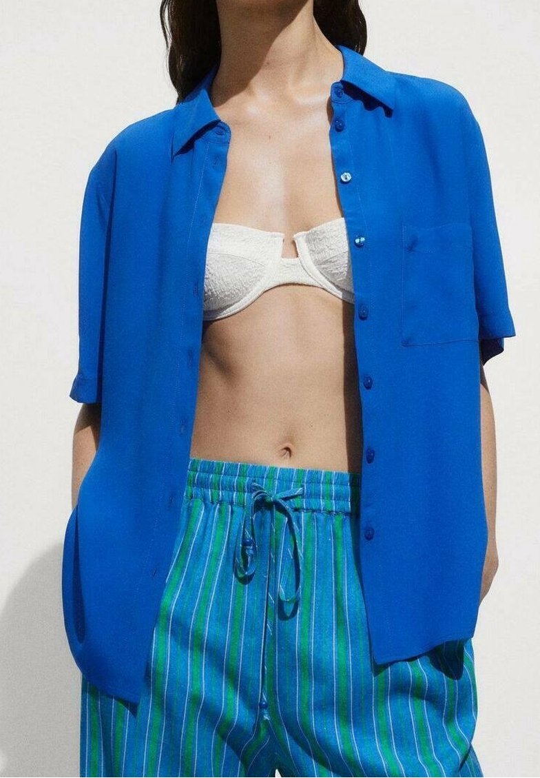Femme portant une chemise bleu vif à manches courtes ouverte, un soutien-gorge bandeau blanc et un pantalon rayé bleu-vert à cordon, debout devant un fond uni.