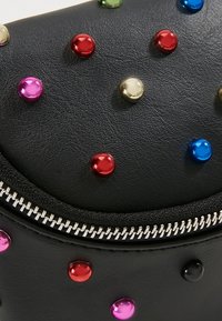 Surface en cuir noir avec des clous métalliques ronds multicolores et détail d'une fermeture éclair argentée courant horizontalement.