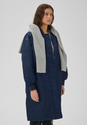Femme portant un long manteau bleu marine avec un pull gris clair posé sur ses épaules, debout devant un fond clair uni.