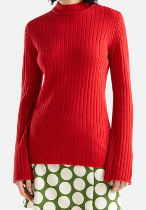 Tæt siddende rød ribstrikket sweater med lange ærmer kombineret med grøn nederdel med store hvide polkaprikker, vist på en stående person.