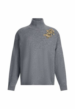 Maglione grigio a collo alto con maniche lunghe e un diadema decorativo di gioielli d'oro e trasparenti sulla spalla sinistra.