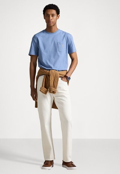 Tricou din bumbac de culoare albastru deschis cu un buzunar în față, pantaloni de culoare crem și un pulover împletit din lână maro, legat în jurul taliei; pantofi maro.