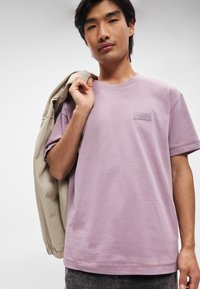 T-shirt lilla a maniche corte realizzato in materiale morbido, con scollatura a girocollo e una piccola etichetta a patch sul petto. Il modello tiene una giacca beige.