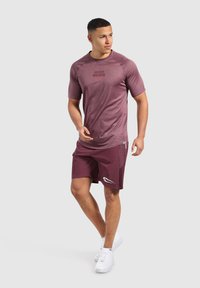 Bordový sportovní triko s krátkým rukávem a logem, spárované s bordovými šortkami s boční kapsou a bílými akcenty. Bílé tenisky.