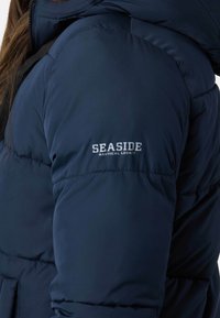 Marineblå pufferjakke lavet af isoleret stof. Har syede sektioner, en lynlåslukning og et logo med teksten "SEASIDE" på skulderen.