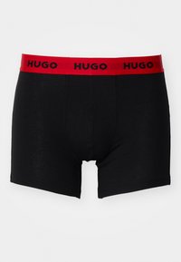 Black boxer briefs with a red elastic waistband featuring the repeated brand name "HUGO" in black lettering. Smooth fabric, mid-thigh length.