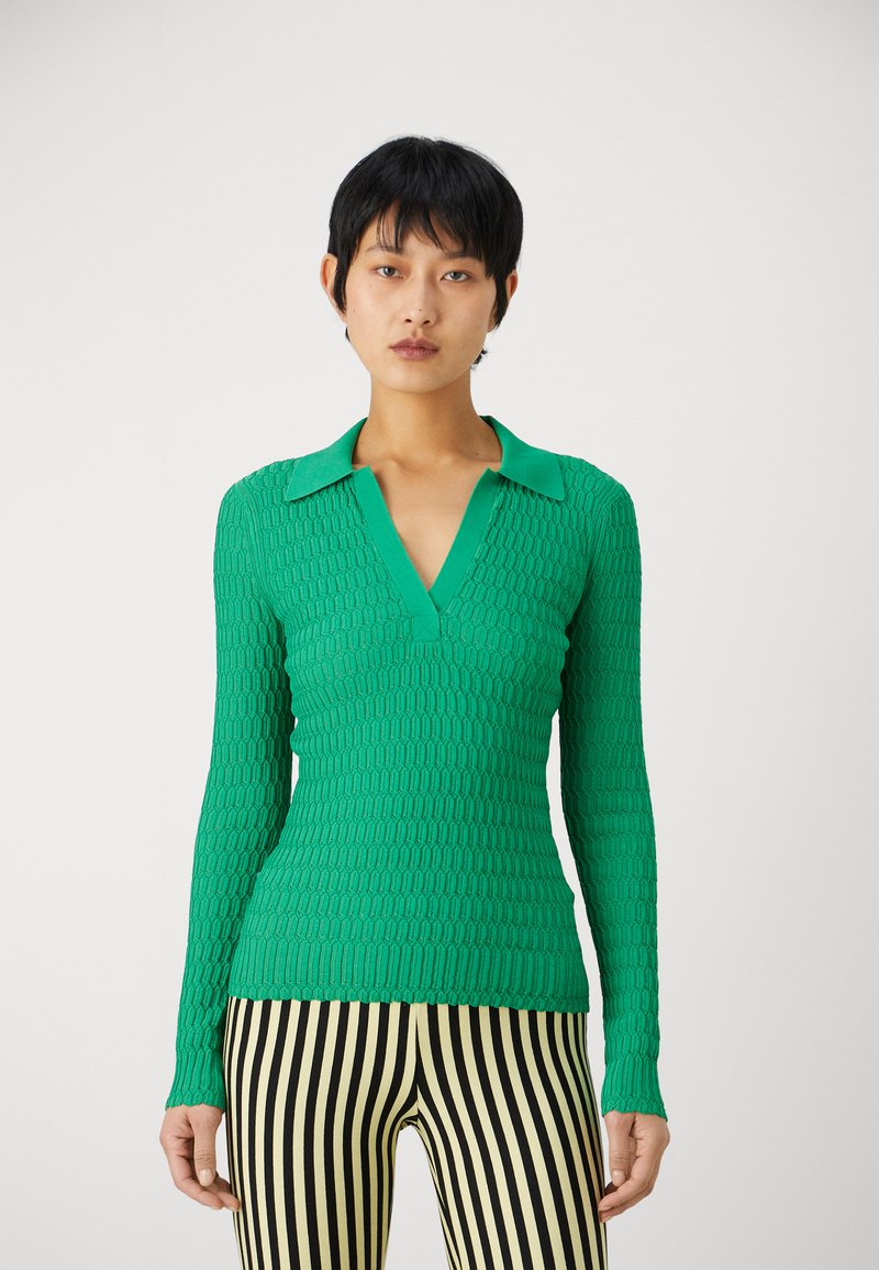 Bluză verde cu mânecă lungă, cu un model texturat, de tip V și cu guler. Asortată cu pantaloni raiți negri și galbeni.