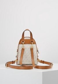 Vaaleanruskea ja kerma kuvioitu minireppu, jossa on ruskeat nahkahihnat, kultaiset yksityiskohdat ja vetoketjukiinnitys yksinkertaisella taustalla. Rakenteellinen materiaali.