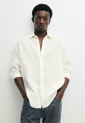 Junger Mann mit hochgekrempeltem langärmligem weißen Leinenhemd und blauen Jeans, steht mit den Händen in den Taschen vor einer schlichten weißen Wand.