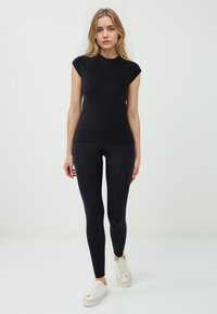 Donna in un top nero aderente e leggings, in piedi di fronte a uno sfondo bianco semplice, con scarpe da ginnastica bianche. I capelli sono sciolti e ondulati.