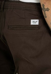 Braune Cordshorts mit einer Gesäßtasche, die ein weißes Label mit der Aufschrift "Reell est. 1997" trägt. Strukturiert Oberfläche, schlichtes Design.