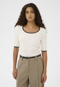 Bluza albă cu dungi, cu mâneci scurte și margine neagră în jurul decolteului rotund, asortată cu pantaloni bej cu talie înaltă și curea neagră.