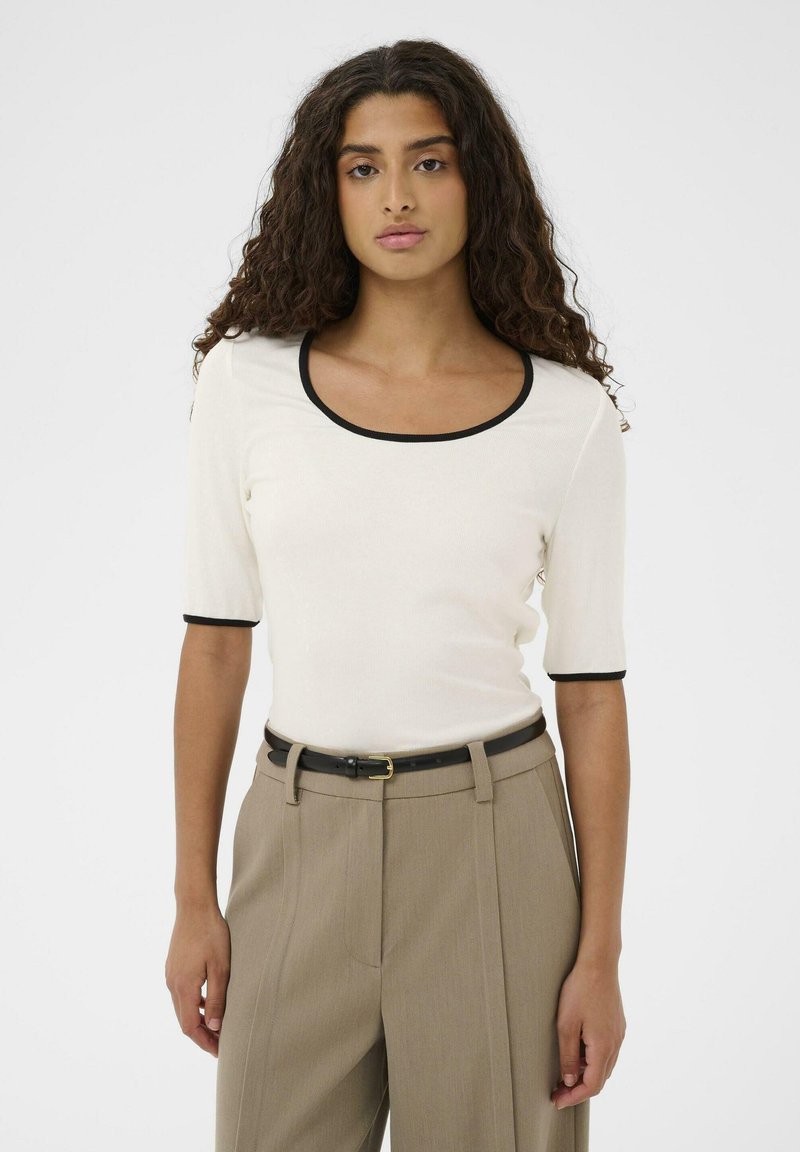 Bluza albă cu dungi, cu mâneci scurte și margine neagră în jurul decolteului rotund, asortată cu pantaloni bej cu talie înaltă și curea neagră.