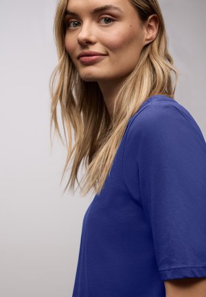 Femme blonde à la peau claire portant une chemise bleue à manches courtes, regardant légèrement de côté avec une expression neutre sur un fond uni.