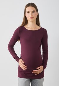 Merlot spalvos ilgos rankovės marškiniai su plačiu apvaliu kaklu, minkšta tampri medžiaga ir prigludusi forma, dėvimi su rankomis ant pilvo, derinami su pilkais džinsais.