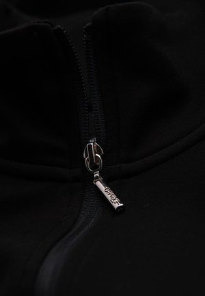 Svart zip-up jacka med hög krage, med en metalldragkedja med ingraverad detalj. Materialet har en jämn, mjuk textur.