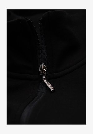 Zwarte zip-up jas met een hoge kraag, voorzien van een metalen ritssluiting met gegraveerde details. Het materiaal heeft een gladde, zachte textuur.