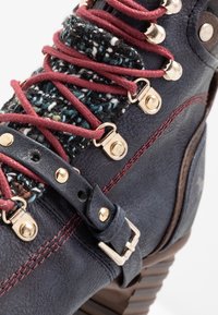Bota de cuero navy con cordones rojos, ojales dorados, una lengüeta de tela texturizada multicolor, costuras rojas y una correa ajustable con hebilla.