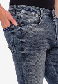 Jeans en denim de couleur indigo dégradé avec un motif texturé. Comprend des poches avant, une fermeture éclair et des patchs logo sur la ceinture.