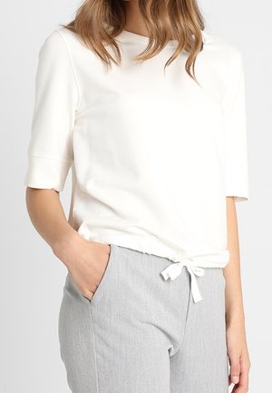 Vrouw met een wit topje met korte mouwen en een knoop aan de voorkant, en lichte grijze broek met hoge taille, hand in zak, staand tegen een effen achtergrond.