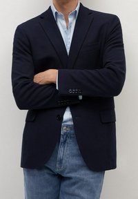 Granatowa marynarka o teksturowanej strukturze z klapami, przednimi kieszeniami i guzikami przy mankietach, w połączeniu z jasnoniebieską koszulą i dżinsami.