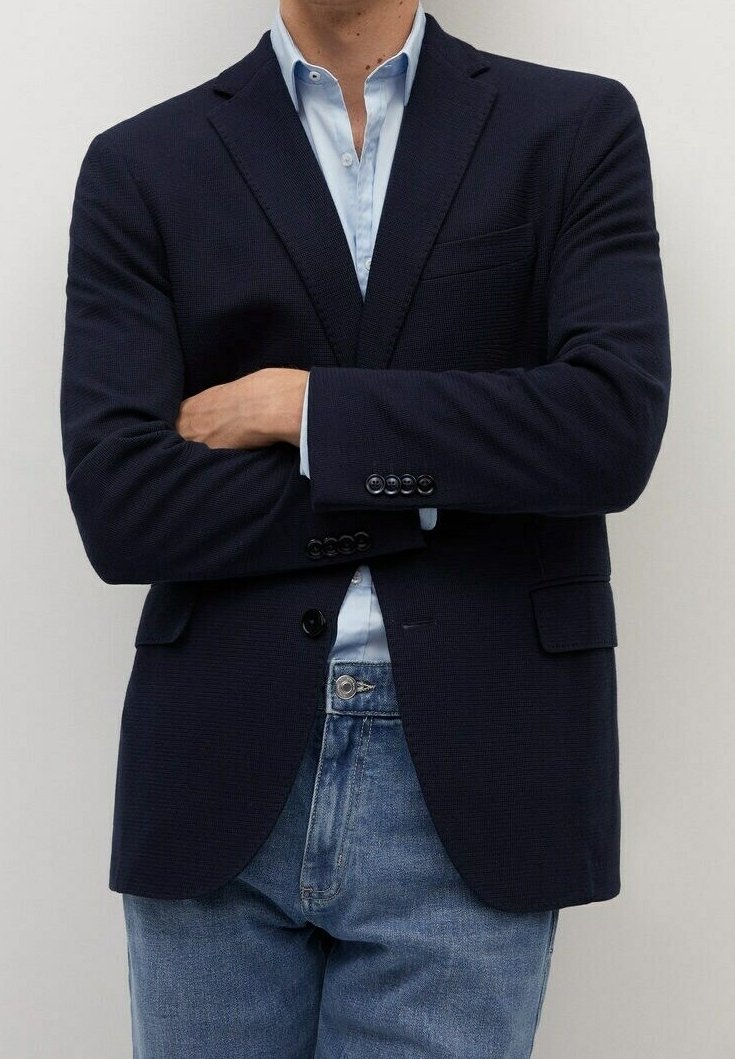 Granatowa marynarka o teksturowanej strukturze z klapami, przednimi kieszeniami i guzikami przy mankietach, w połączeniu z jasnoniebieską koszulą i dżinsami.