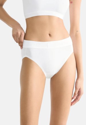 Donna che indossa slip bianchi in cotone a vita alta e un top corto bianco, che sistema la vita con la mano sinistra davanti a uno sfondo semplice.