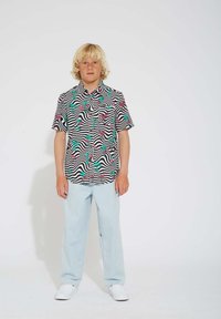 Chemise à manches courtes avec un motif ondulé noir et blanc, ornée de flamants roses et de feuilles de palmier vertes ; associée à un jean bleu clair et des chaussures blanches.