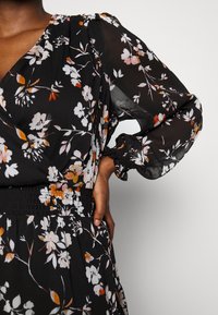 Robe fleurie noire en tissu transparent, ornée de fleurs blanches et orange, manches bouffantes et taille smockée.
