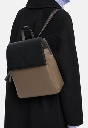Osoba ubrana w czarny płaszcz z dwukolorowym plecakiem, posiadającym czarną klapę i taupe kolor korpusu, noszonym na jednym ramieniu.