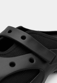 Chaussure noire avec une tige en tissu texturé, une large bride et des trous de ventilation, conçue pour le confort et la respirabilité.