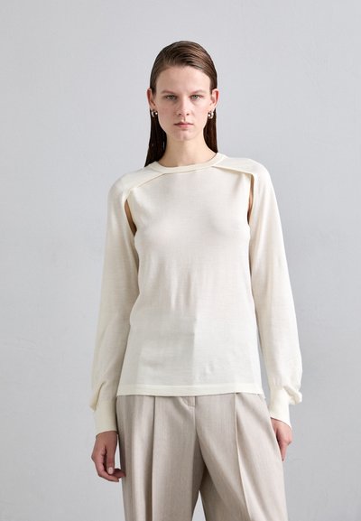 Haut à manches longues en crème clair, en tissu doux et texturé ; doté d'un détail de découpe unique aux épaules et de poignets côtelés. Associé à un pantalon beige.