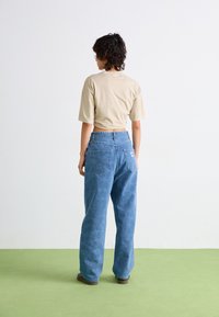 Beige cropped t-shirt, loszittende lichtblauwe spijkerbroek. De spijkerbroek heeft twee achterzakken en een zichtbaar merkteken. Groene vloer achtergrond.