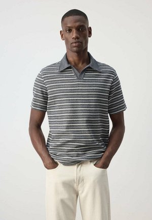 Hombre con una camisa polo de punto de rayas grises y blancas de manga corta y pantalones beige, de pie con las manos en los bolsillos sobre un fondo liso.