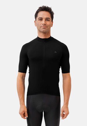 Uomo in piedi rivolto in avanti che indossa una maglia da ciclismo nera a maniche corte aderente e pantaloncini da ciclismo imbottiti neri su sfondo bianco.