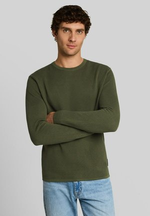 Jonge man met krullend haar, gekleed in een olijfgroen gestructureerd shirt met lange mouwen en lichtblauwe spijkerbroeken, staand met gekruiste armen.