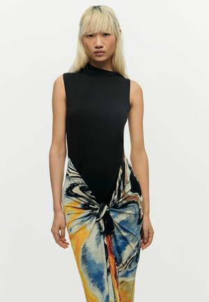 Jonge vrouw met lang blond haar draagt een mouwloos zwart topje en een kleurrijke, abstracte, patroonrok die vooraan is gestrikt, tegen een witte achtergrond.