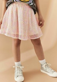 Una falda de color rosa claro, hasta la rodilla, con lentejuelas iridiscentes, que presenta un diseño floral texturizado y una cinturilla elástica. Combinada con zapatillas plateadas.