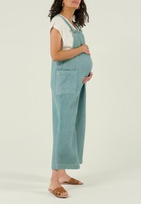 Zwangere vrouw in blauwe textuuroveralls en wit shirt staat zijwaarts, haar handen voorzichtig haar buik steunend, met bruine slippers aan.