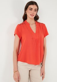Blusa rossa a maniche corte con scollatura a V, dettagli in pizzo sulle spalle e parte anteriore plissettata. Realizzata in tessuto leggero per il massimo comfort.
